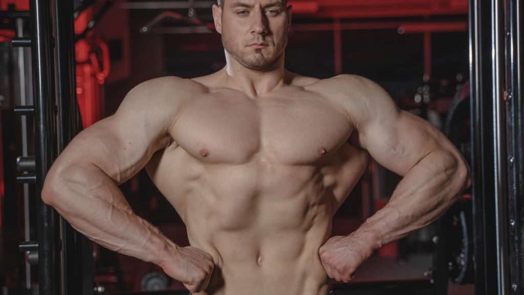 Intense shot of a muscular man posing topless in a gym setting showcasing physique.