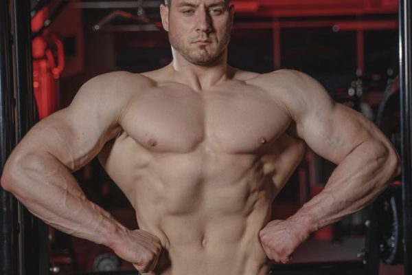 Intense shot of a muscular man posing topless in a gym setting showcasing physique.